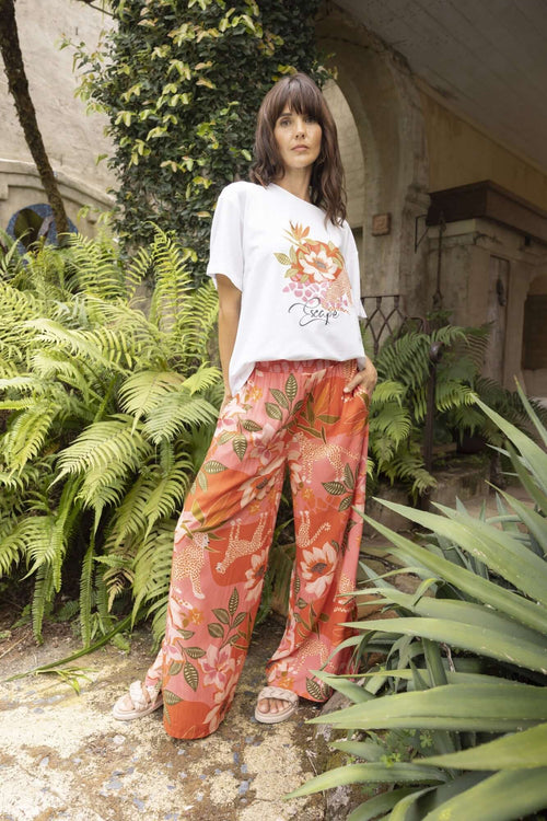 Model wearing Aria Pants with a floral print, standing among lush greenery, showcasing summer style.