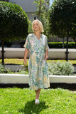 A woman wearing the Kazia Dress, a cream midi dress with green and yellow patterns, smiling in a sunny garden.