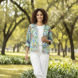 A woman wearing the Sante Fe Jacket 2 in a park, paired with white pants and sandals, surrounded by greenery.