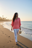 Woman in a coral Barika V neck top walking on the beach at sunset, embodying elegant summer style.