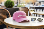 Joy Caps in pink with Prosecco embroidery on a table beside a glass of wine and sunglasses.