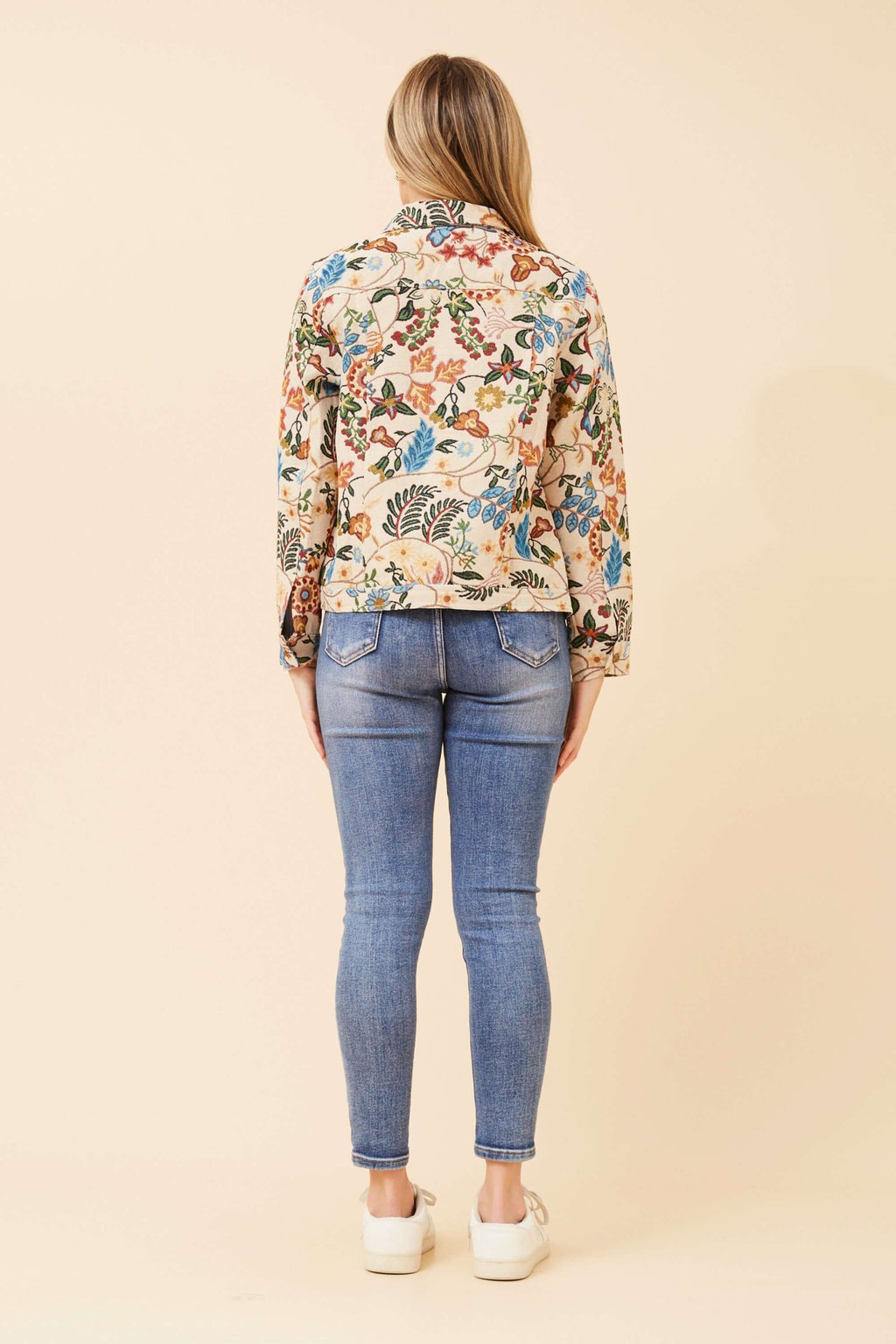Back view of a woman wearing the CKM Tapestry Jacket with floral patterns, paired with blue jeans and white shoes.