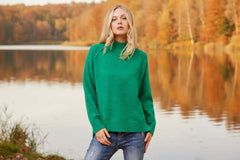 Model wearing the Katya High neck jumper in vibrant green near a serene lake surrounded by autumn foliage.