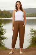 Model wearing Roma Linen Pants with a white tank top and sandals against a warm orange background.