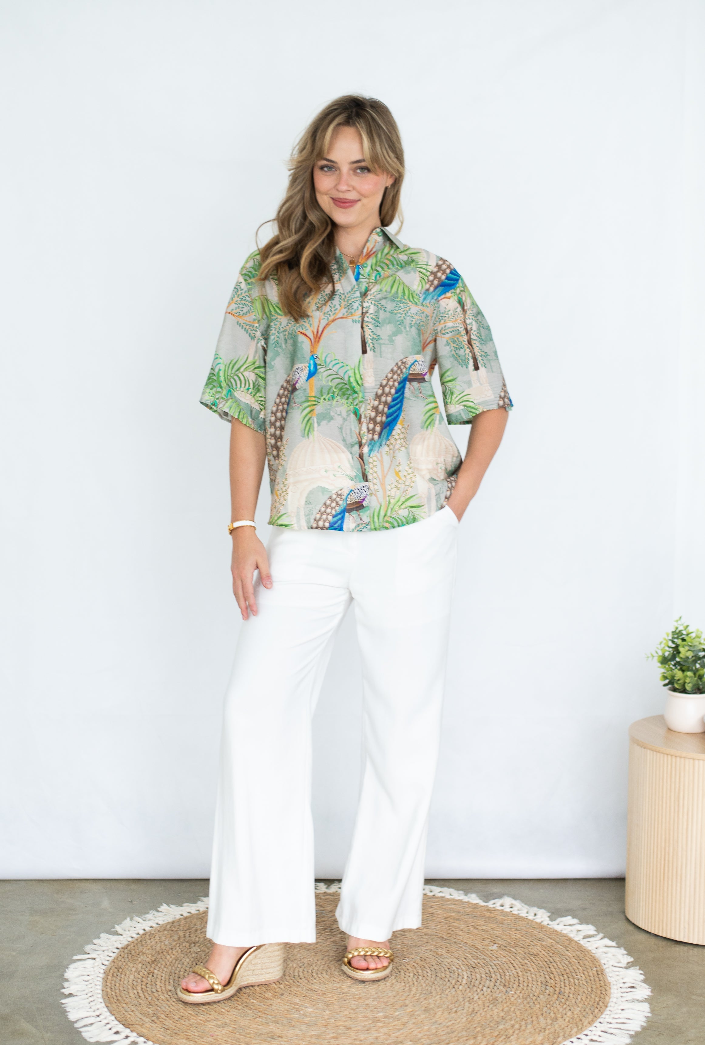 Model wearing the Peacock in Palace shirt with vibrant peacock print and white pants against a neutral backdrop.