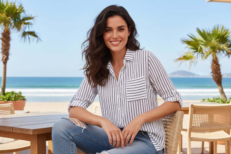 Alma Striped Shirt worn by a model at a beachside table with palm trees in the background.