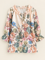 A long-sleeve blouse with a multicolor floral print on a cream background, featuring ruffled trim at the neckline and cuffs, displayed against a solid light beige background.