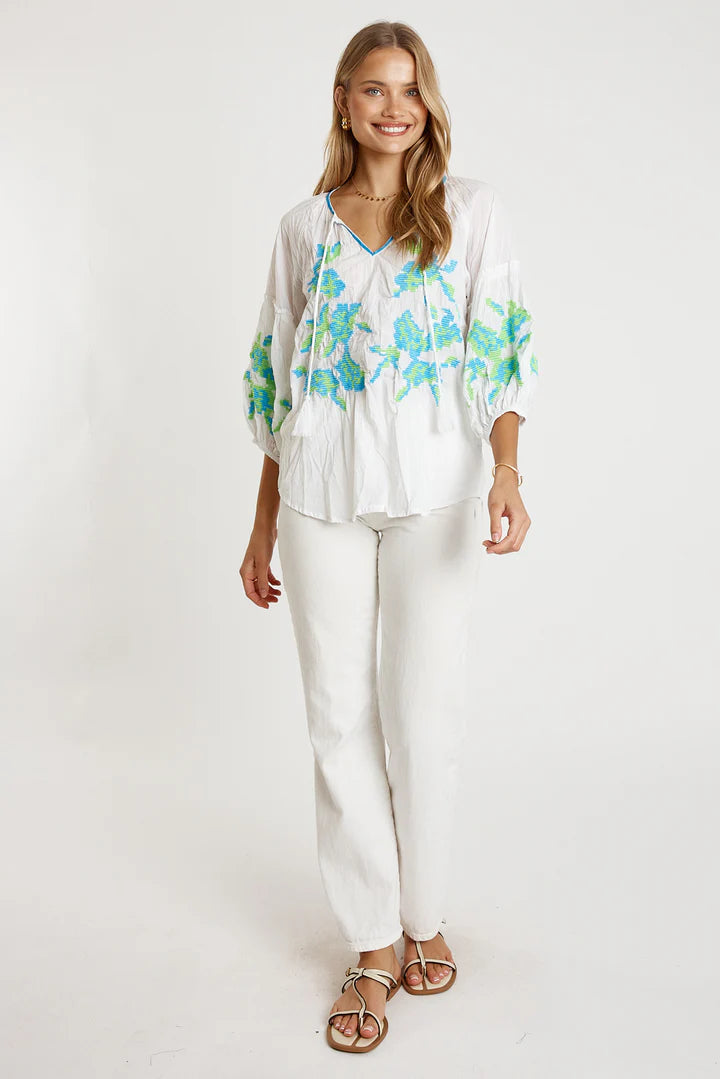 Woman in LulaLife Rosalind Top White with blue and green embroidery, paired with white trousers for a stylish summer look.