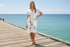 A woman in the Sahara Dress White stands on a wooden pier by the ocean, showcasing its boho charm and embroidered details.