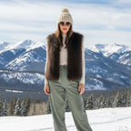 Model wearing a Mongolian Vest with faux Afghan fur, paired with a knit hat and sunglasses amid snowy mountains.