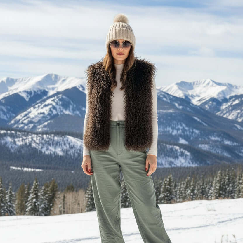 Model wearing a Mongolian Vest with faux Afghan fur, paired with a knit hat and sunglasses amid snowy mountains.