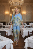 A model wearing the Silk Scenic Pants Sky paired with a matching top, exuding elegance in a luxurious dining setting.